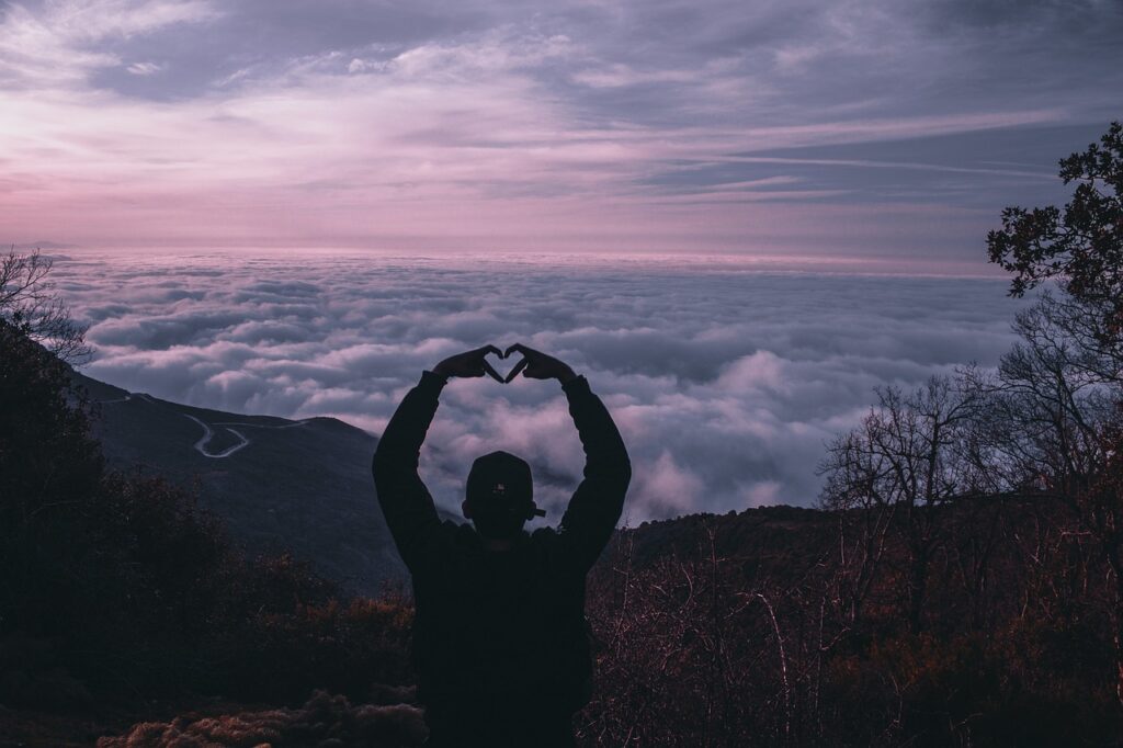 Man on a mountain with his hands over his head while his hands make a heart shape.